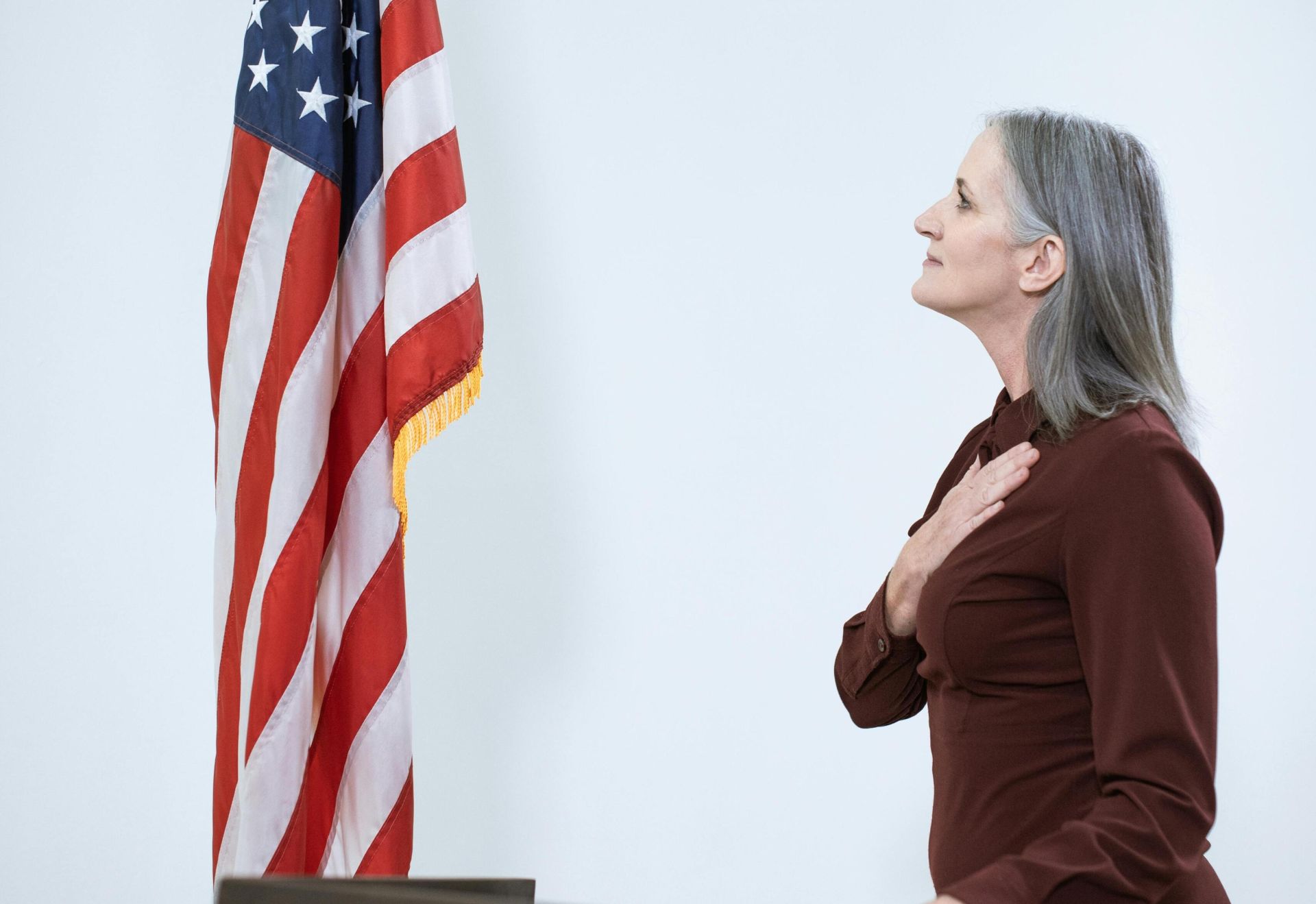 A senior woman pledges allegiance at a podium next to an American flag.