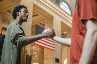Two young men exchange a friendly handshake indoors with an American flag in the background.