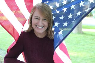 A woman smiling warmly while holding an American flag outside on a sunny day.